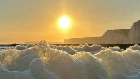 Grazyna Hammond  A close of sea foam with the sun and cliffs looming in the background 