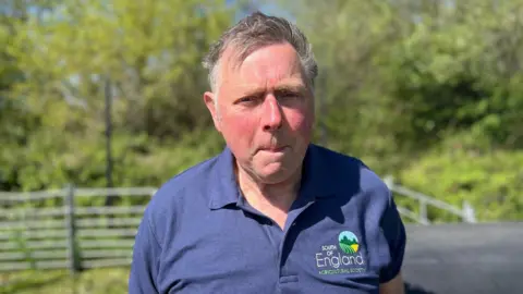 BBC/Sam Dixon-French An older man with a blue polo shirt looks at the camera with a sheep pen in the background.