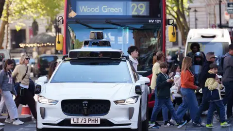 A white self-driving Jaguar vehicle fitted with roof-mounted sensors driving in central London, flanked by pedestrians crossing the street and a red double-decker bus directly behind it.