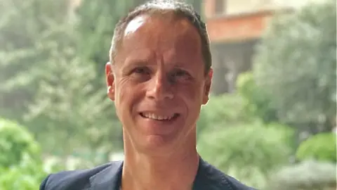 Jersey Opera House The picture shows a person standing outdoors in what appears to be a historic or garden setting. The background includes tall green trees, shrubs, and an old stone building with arched windows, suggesting a courtyard or heritage site. 