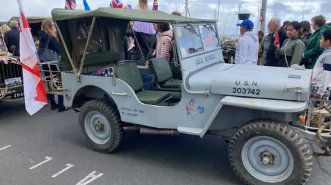 A US military jeep which has a canvas roof. It is pale grey and has no doors. Printed on the side is OFFICIAL USE ONLY NO CIVILIANS. On the bonnet is printed USN 203742 and under the windscreen reads USS NEVADA. There are spectators surrounding it.