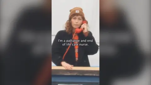 A woman holds a red telephone receiver on a bridge. She has red hair and wears a beanie hat and black coat