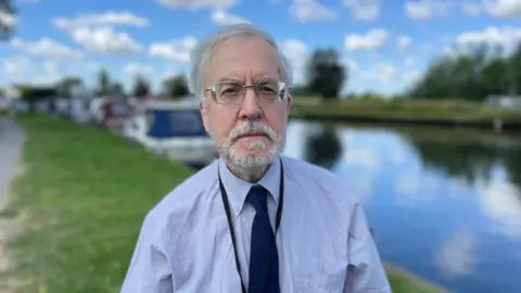 Shaun Whitmore/BBC Barry Darch, wearing a pale blue shirt and navy tie, stands on the banks of a river with moored boats in the blurred background. He has grey hair and beard and glasses.