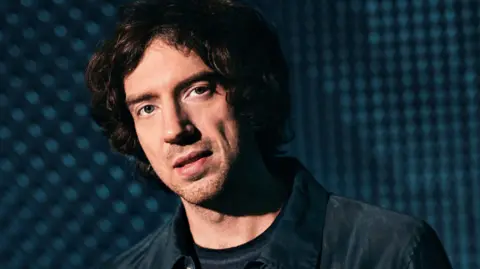 Gary Lightbody in dark jacket with blue, blurry background. He has long-ish dark hair and some stubble