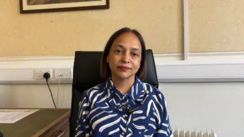 Balvinder is wearing a blue and white patterned top and she is sat in a chair in an officer. She has sort bobbed hair.
