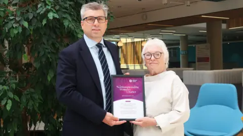 A man and a woman are holding a framed certificate. The man is wearing a suit and the woman is wearing a white top. There is a blue chair behind them and a large indoor plant 