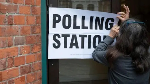 A woman sticks up a large sign reading "polling station" on the door of a church hall. 