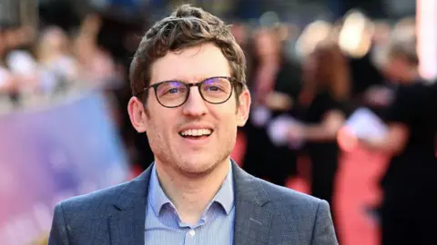 Elis James has brown hair, black glasses and is smiling. He is wearing a blue blazer and blue, striped shirt