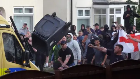 Men, some wearing masks and holding an England flag, throw a wheelie bin at a police van