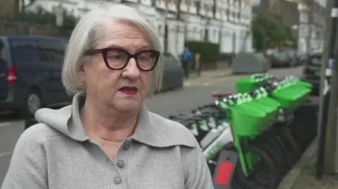 A woman with grey hair and glasses standing on the pavement in a residential street, with a row of bright green e-bikes in the background behind her.