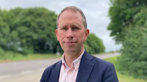 Matthew Barber has short brown hair with specks of white. He is wearing a dark suit jacket over a light pink shirt and is stood in front of a country road and bushes that are blurred slightly by the perspective of the shot.