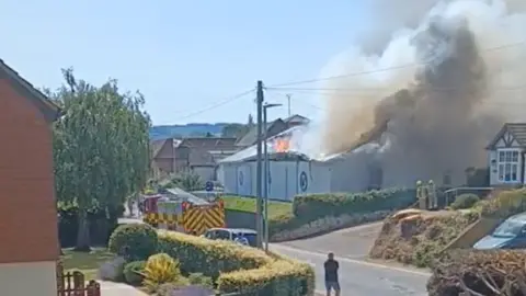 Chris Hunt A large amount of smoke and flames billow out of the gutted roof of a social club as a fire engine drives past. There are houses surrounding the social club and a pedestrian can be seen watching on from the pavement.