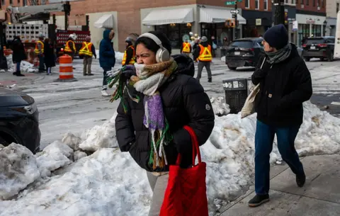 Getty Images A woman is pictured screwing her face with a scarf tied around her mouth, and the hands in the pockets of her puffer jacket as she walks down a NYC street. Piles of shovelled snow are seen in the background