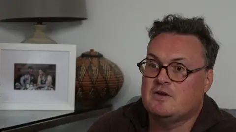 A man wearing black-rimmed glasses looks towards the camera. A picture of his wife and children can be seen in the background