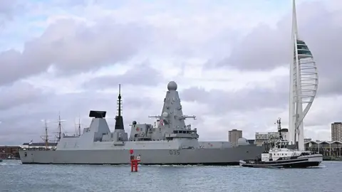 HMS Dragon, a large grey destroyer ship, sits on the water in Portsmouth Harbour, under a cloudy sky.