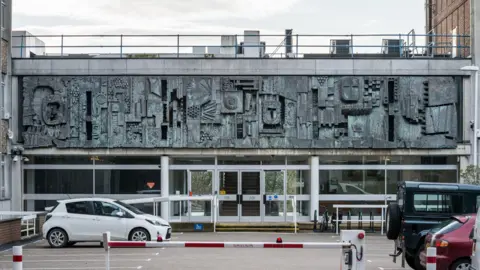 Getty Images A picture of the relief mural by William Mitchell, on the wall of County Hall, Saint Anne's Crescent, Lewes, East Sussex, 2014, a grey and white building created in 1967. Three vehicles are parked in front of the main entrance and the car park barrier is down.
