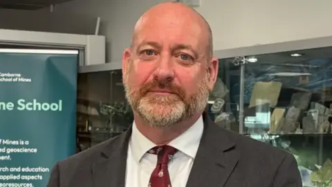 Perran Moon has no hair and has a grey/ginger beard. He is wearing a dark jacket and a red tie, and a white shirt.