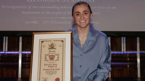 Women in a blue blouse with brown hair wearing a necklace. She is smiling and holding a certificate declaring her 'Freedom of the Town' award.