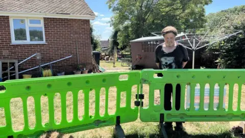 This image shows a backyard scene with a person standing behind a green plastic barrier. In the background, there's a brick house with white-framed windows, a wooden shed, and various garden tools and pots scattered around. A clothesline is also visible. The man is looking down into the hole which is in front of the barrier with some concern.