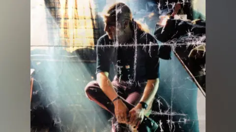 Contributed A scan of a physical photograph that shows a man sitting in a chair with one leg crossed over the other. He is looking to the left. He has long light coloured hair which has been tied back. He is wearing a dark top with sleeves that go to his elbows and red trousers.