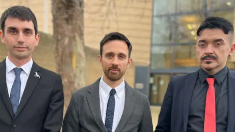 Three men lined up outside Cambridge Crown Court, all wearing jackets shirts and ties. Daniel Kidby has short dark brown hair; Ben Newman has short lighter brown hair and a close cropped beard; Beau Sebastian has a moustache and goatee.
