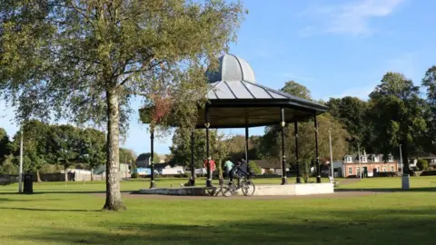 A bandstand in green parkland. There are a few trees and some children playing on their bikes. 