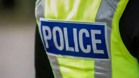 Generic shot of a police officer wearing a hi-vis yellow jacket with the word "police" printed on the back in white letters on a blue background