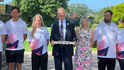 BBC Group of people stand outside a gated entrance in a garden setting; several wear white “King’s Baton Relay Glasgow 2026” shirts while a central figure in a suit holds a decorated baton, with greenery, trees, and a large open gate behind them.
