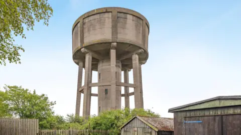 The water tower which is a round, concrete structure towering above the treeline and some wooden sheds. It has a central column down the centre with windows and about seven slimmer legs around it. It has a large, round top which is windowless. The sky above is blue and cloudless.