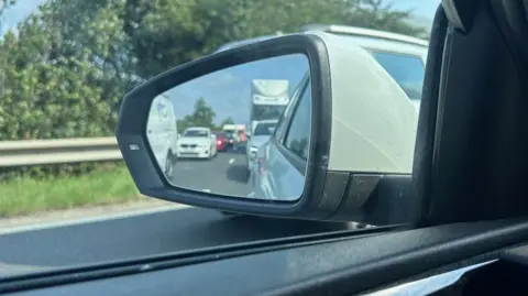 BBC A shot of queuing traffic taken through the passenger wing mirror of a car. There is a long line of vehicles, including lorries, cars and vans.
