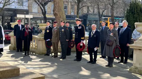 Men and women in military uniform, some holding poppy wreaths, gather to pay their respects at a war parade. An old man is standing at a microphone, talking.