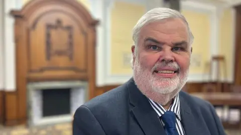Dave Thomas is wearing a blue and white striped shirt and a navy blue tie and jacket and is standing in a committee room