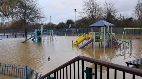 Flood water covers the entirety of a children's play area with the water level up to the top of the children's swings. Two pieces of climbing equipment are also submerged as is a picnic bench in the right of the picture.