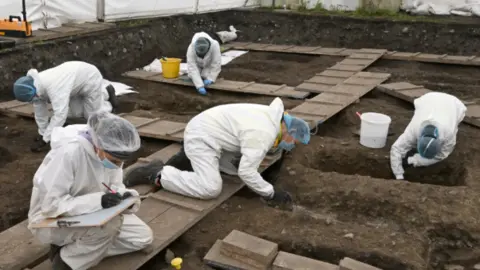 Five forensic staff wearing white hazmat suits, plastic hair nets and blue medical masks work under a large tented enclosure during the excavation. They are kneeling on wooden gangways in a large pit and leaning into shallow graves-like hollows, scraping at the soil. Plastic buckets are placed beside some of the workers. One worker is writing on a clipboard. 