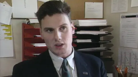 A young man wearing a suit and tie sits in an office in front of paperwork