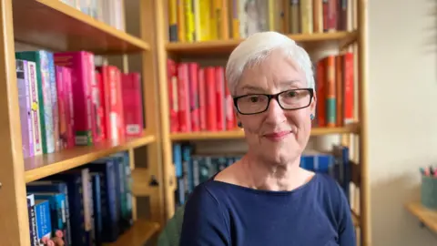 BBC Alex Morgan sat in front of a bookcase in her home. She has short white hair and blue eyes. She is wearing glasses with a square dark frame. She is wearing a blue top. The books behind her are on a wooden bookcase, organised by colour.