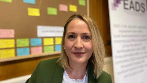 Laura Coffey/BBC Julia Green, a woman with blonde hair wearing a green jacket and white top, looking directly at the camera. She is wearing two gold necklaces and gold earrings. She is sitting in an office with a large cork board behind her with coloured notes.
