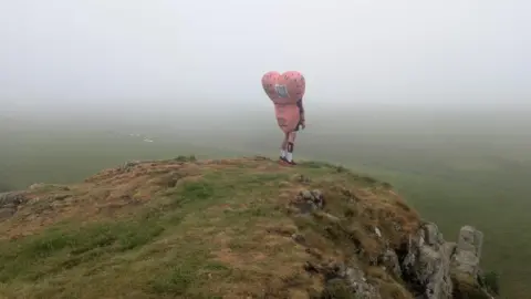 Robin Cancer Trust A man dressed in a large pink costume shaped like testicles stands on top of a foggy hill looking out to the valley below.