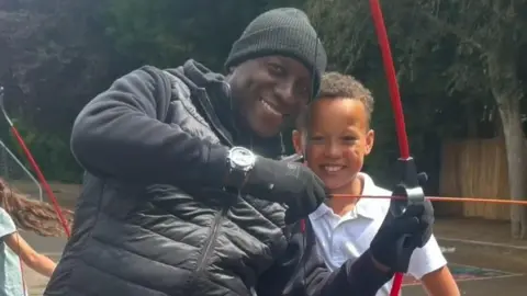Family A man and a boy stand close together. They are both smiling. The man is holding a bow and arrow.