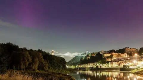 Sam Binding The northern lights are visible over the Clifton Suspension Bridge, with the lights of the bridge and Clifton reflected in the water of the River Avon below. The northern lights are a mixture of purple and green