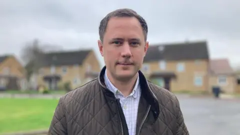 Ashley looks at the camera, wearing a padded brown coat with a check shirt underneath. Blurred behind him is a residential area, with houses and green area.