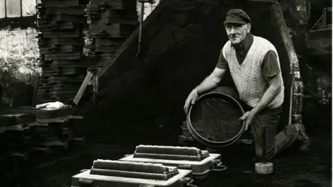 Gary Norman Black and white photo of a man on one knee, holding a round object over a set of foundry moulds. He is looking at the camera and wearing a knitted vest over a short-sleeved shirt and a cloth cap.