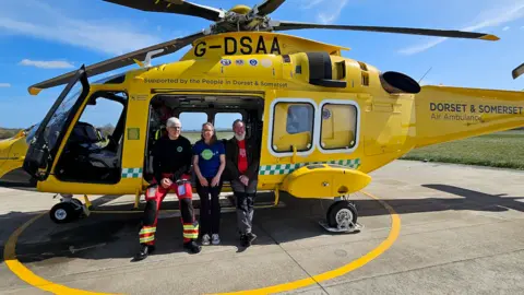 Zoe Langley-Wathen The couple and a member of the air ambulance to their left sitting on the side of an open yellow helicopter. They are all faceing the camera with their hands on their laps and smiling. They are on a concrete surface, with a yellow circle surrounding the helicopter, behind them is green grass.