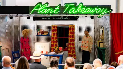 Eden Project The Plant Takeaway is like a small stage holding a kitchen. There is an audience watching it. On the stage are a female and a male mannequin. The female one is wearing a dress printed with aubergines and the male a shirt featuring carrots. There is a table with two glasses and a bowl of fruit and there is a fridge with SMUG printed on it. There is a window with strawberry printed curtains and a mantelpiece.