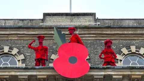 Salisbury City Council The top of a grand building under a grey sky - a huge version of a Royal British Legion poppy is attached to the front and behind it are three soldier-type figures which are covered in knitted poppies.