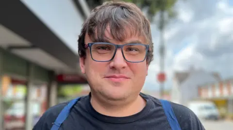 Andrew Simmons has short brown hair and blue-framed glasses. He is wearing a black T-shirt.