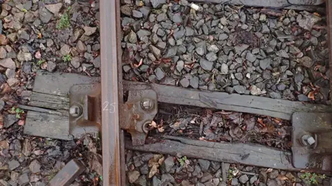RAIB One of the metal train tracks on a gravel covered floor. The wooden sleeper is rotted and askew. The bolt holding the two together has a chunk missing in the bottom right hand corner.