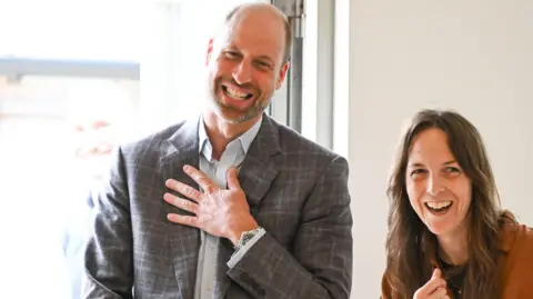 Managing Director Rachel Phillips and the Prince of Wales talk indoors near a bright window with one hand on his chest, both facing toward the camera.

