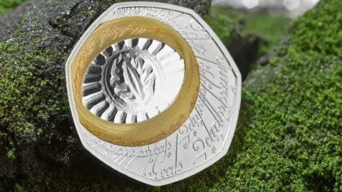 The Royal Mint A single coin on a fake background of granite grey rock frosted in green lichen. the coin is silver with an etched inscription in Elfish, a Lord of the Rings script and language. A gold band, the one ring, also inscribed in Elvish, is at the top left of the seven sided coin. the ring surrounds the eye of sauron etched onto the silver coin with an oval around the eye which is surrounded by a fan of lines to the ring. 
