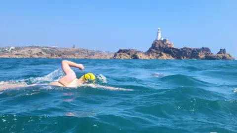 Roger Davies Marie-Helene Sanderson swimming in the sea. She is wearing a purple swimming suit and bright yellow swim hat, A white lighthouse on a rocky headland is in the distance.